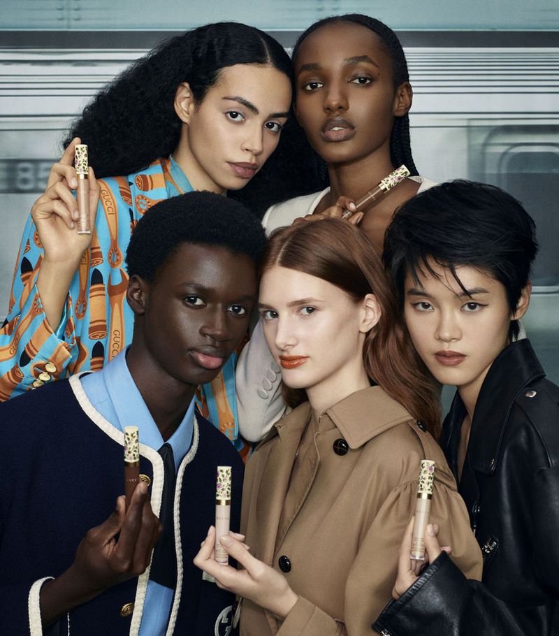 Five diverse models pose together indoors, holding Gucci makeup concealers. They wear stylish jackets and shirts in a beauty product shot.