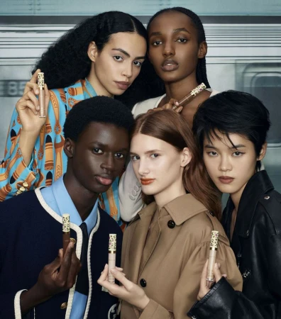 Five diverse models pose together indoors, holding Gucci makeup concealers. They wear stylish jackets and shirts in a beauty product shot.