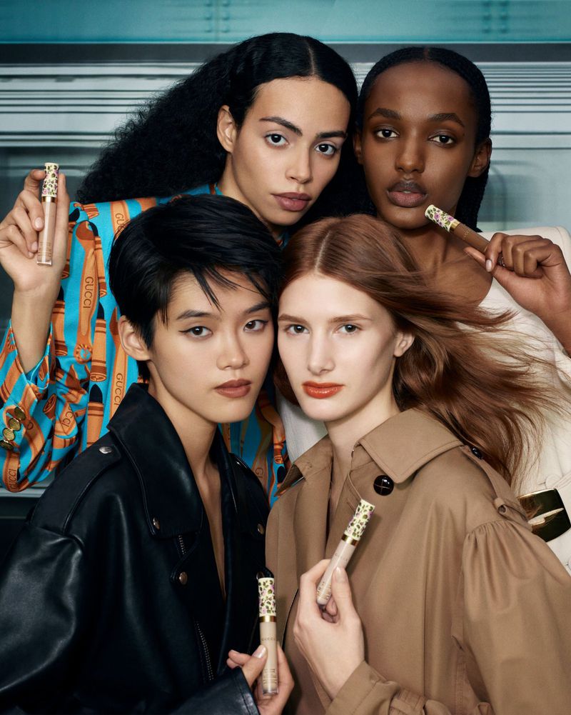 Four diverse models in close-up studio shot, showcasing makeup wands and wearing fashionable clothing against a light blue background.