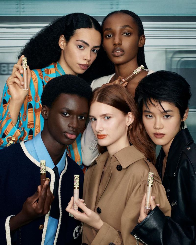 Cosmetics product shot featuring five diverse young adult models holding concealer tubes in a subway setting, promoting beauty and fashion.