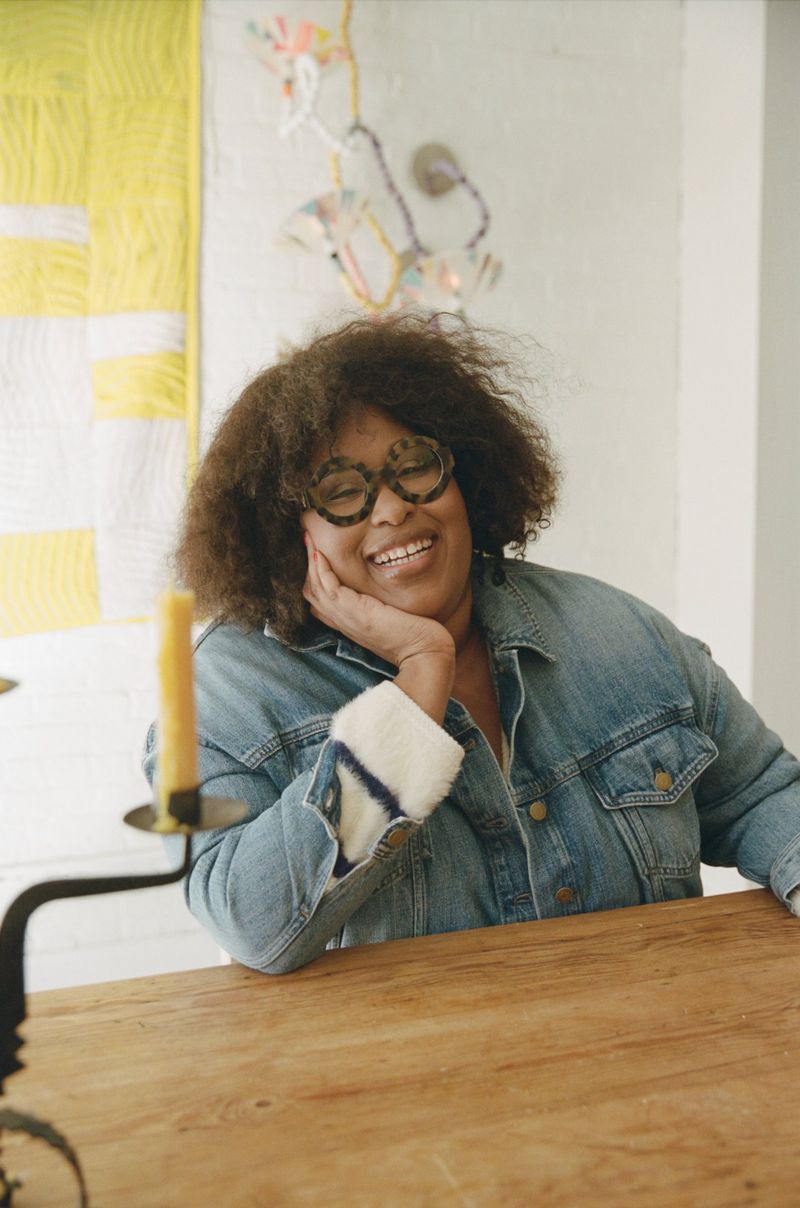 Smiling adult woman with dark curly hair, wearing glasses and a denim jacket, sitting at a wooden table in an indoor setting.