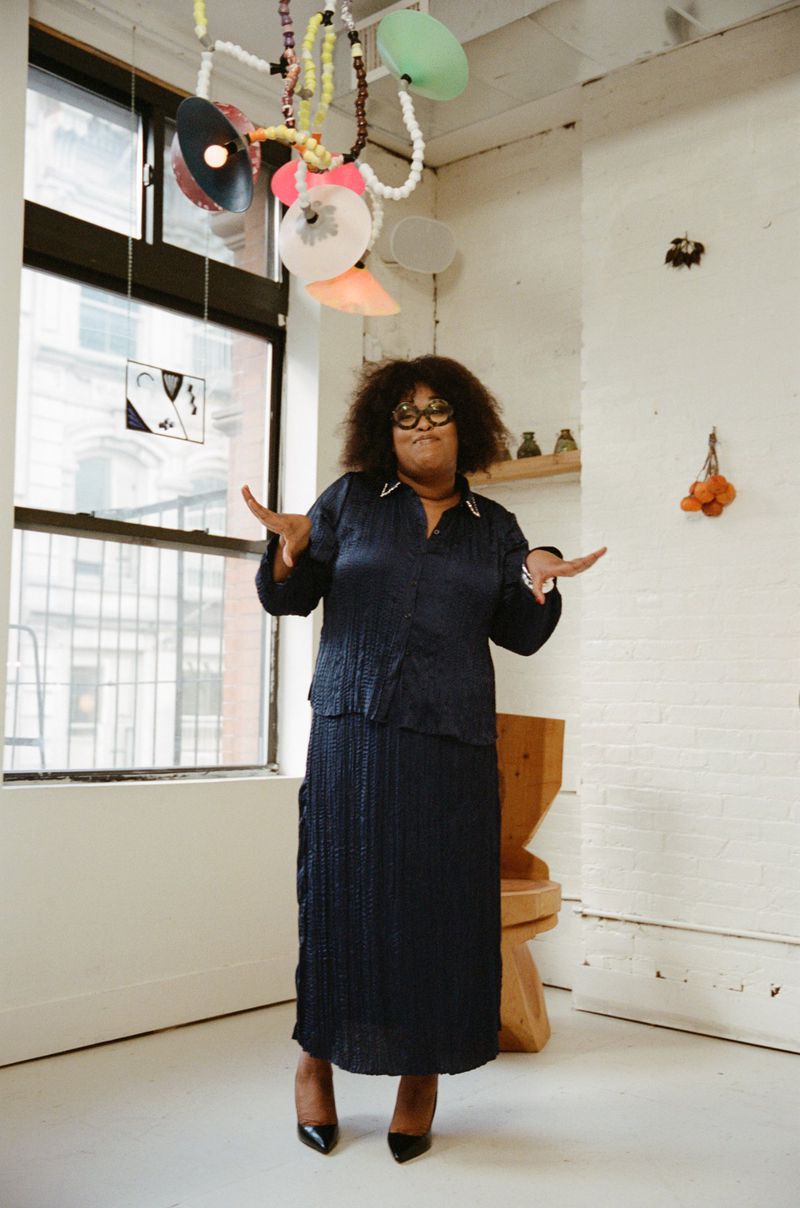 Full shot of an adult female wearing a dark blue suit in an indoor environment with decorative lighting and a wooden stool.