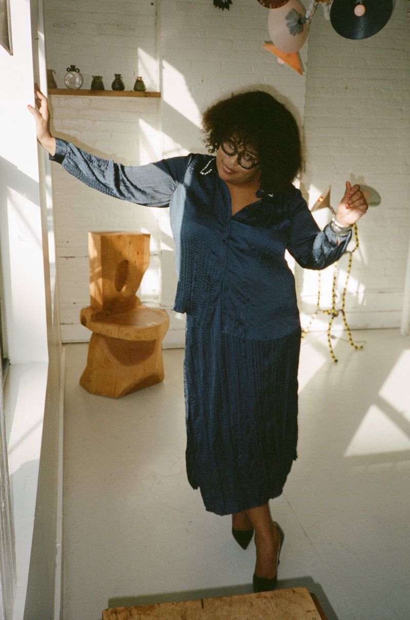 Elegant woman in blue satin dress posing indoors with vintage decor, wearing glasses and jewelry, exudes timeless fashion appeal.