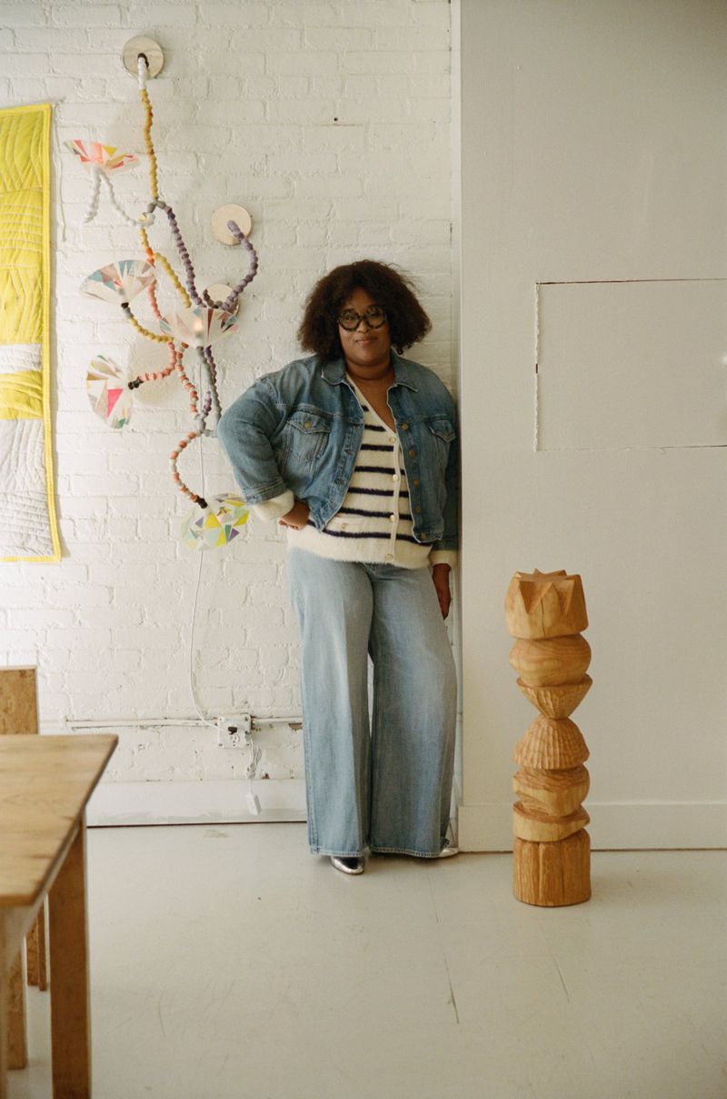 Woman in denim jacket and jeans standing indoors near a wooden sculpture and wall art, posing with hand on hip.