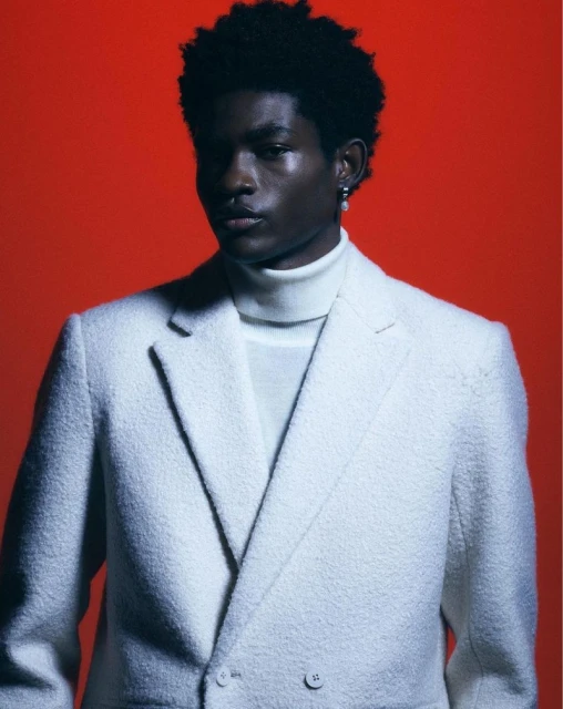 Portrait of a Black male model in a white wool suit and turtleneck against a red backdrop, looking directly at the camera.