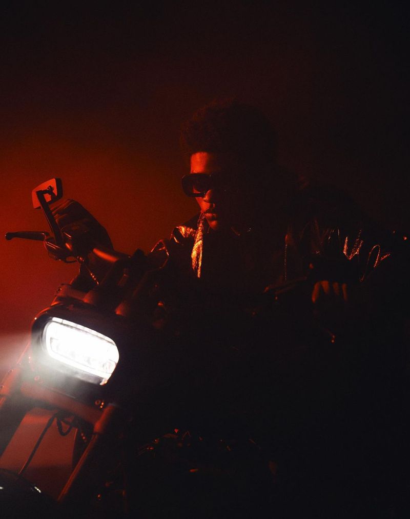 Person riding motorcycle in a dark, dimly lit setting with red tones and bright headlight. The image is dramatic and eye-catching.