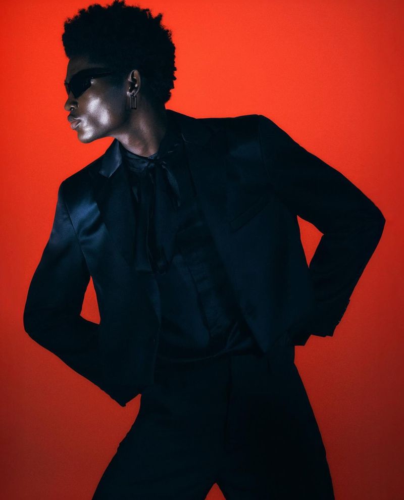 A dark-skinned model poses in a sleek black suit with sunglasses against a vivid orange backdrop, creating a striking and modern aesthetic.