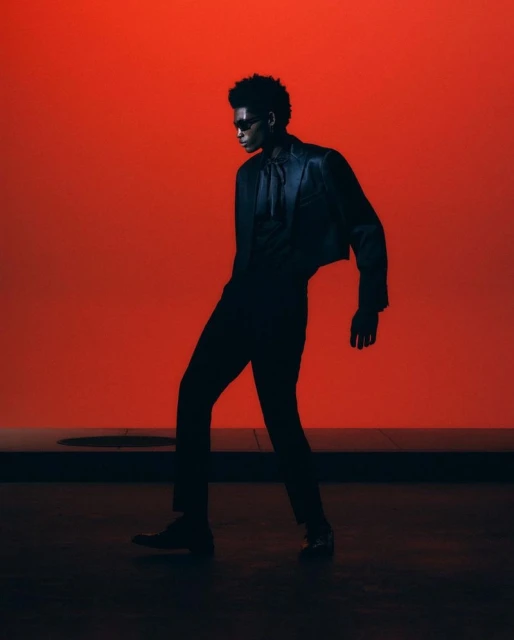 Man in silhouette poses against a vibrant red background, wearing a cropped jacket, dress pants, and sunglasses for a modern fashion statement.