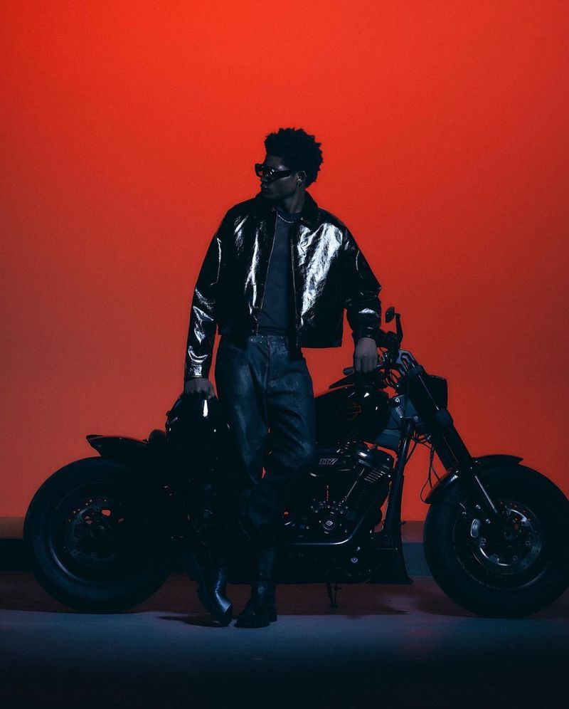 A black man with sunglasses poses with a motorcycle in a studio setting with an orange backdrop, wearing a shiny black jacket and jeans.