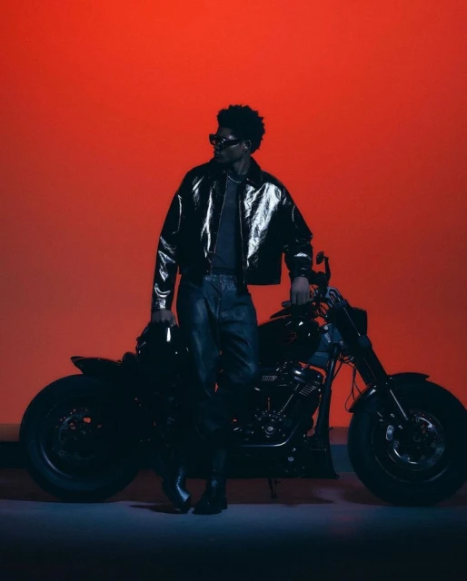 A black man with sunglasses poses with a motorcycle in a studio setting with an orange backdrop, wearing a shiny black jacket and jeans.
