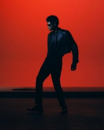 Model Evans Ikechukwu in a black cropped suit and dark sunglasses posing against a solid red studio background.