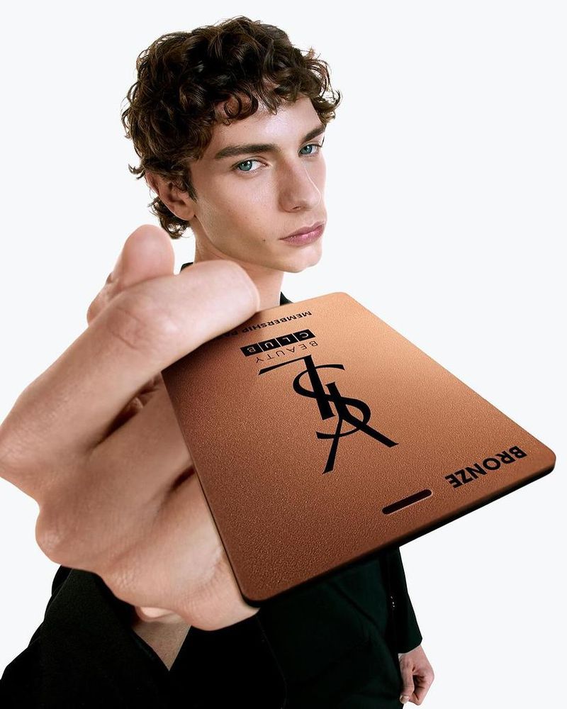 A fashionable young man poses with an Yves Saint Laurent (YSL) Beauty Club Bronze membership card in a close-up studio shot on white.