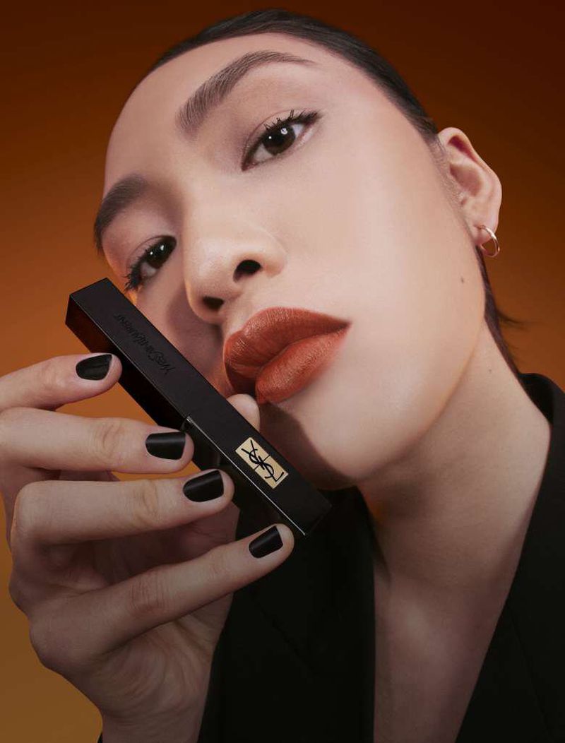 Close-up of an Asian woman's face with a matte orange lipstick holding a black and gold lipstick tube on an orange background.
