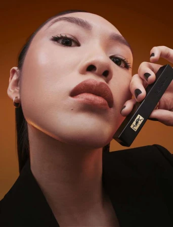 An Asian woman with fair skin and bronze lipstick holds a black YSL lipstick tube against a dark orange backdrop, looking directly at the camera.