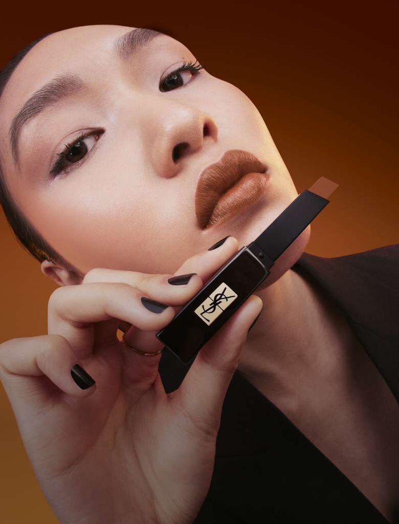 Close-up of an Asian woman with brown lipstick, holding YSL lipstick in hand. The beauty shot is set against a gradient brown backdrop.