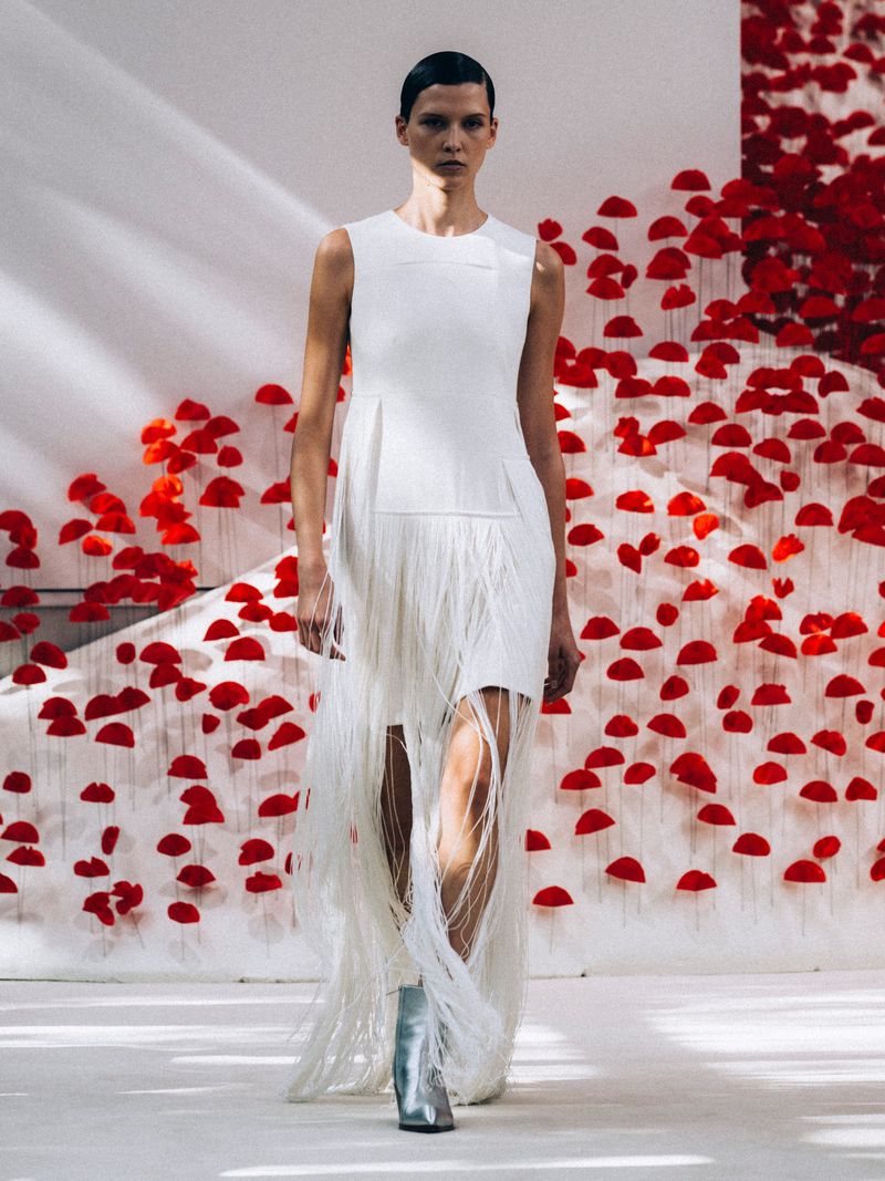 A fashion model walks a runway in a white dress with a fringe skirt and silver boots in front of a red floral display.