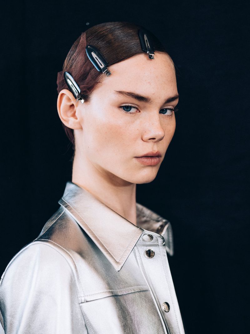 Portrait of a young adult woman with hair clips in her short brunette hair, wearing a silver jacket, against a dark background.