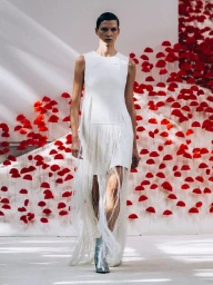 Model walking on a runway in a sleeveless white dress with floor-length fringe and silver boots against a red floral backdrop.