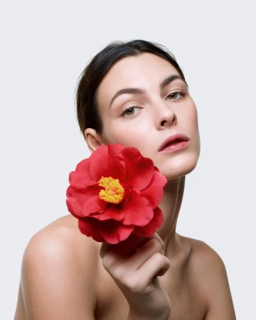 Portrait of a light-skinned woman holding a red camellia flower against a neutral background in a beauty or fashion context.
