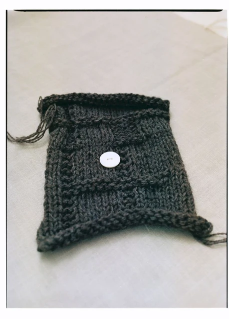 Knitted dark grey bag with a white button closure, showcasing craftsmanship and design on a neutral backdrop.