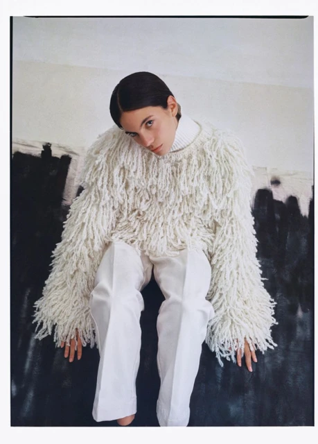 A woman with dark hair in a fuzzy sweater and white pants sits indoors against an abstract backdrop in a portrait-style fashion photograph.