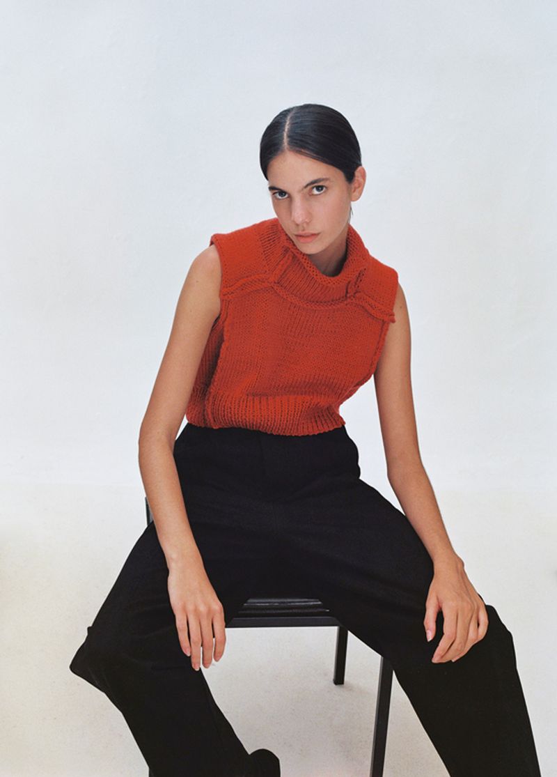 Model wearing a rust-colored knit sleeveless turtleneck top and black pants seated on a chair against a plain white background.