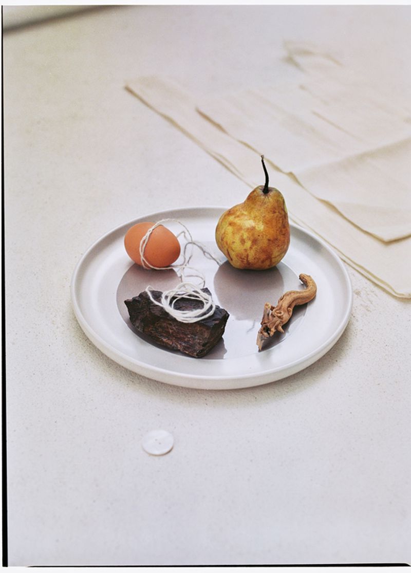 Still life of a pear, egg, stone, and other objects arranged on a plate against a minimalist white background, soft, diffused lighting.
