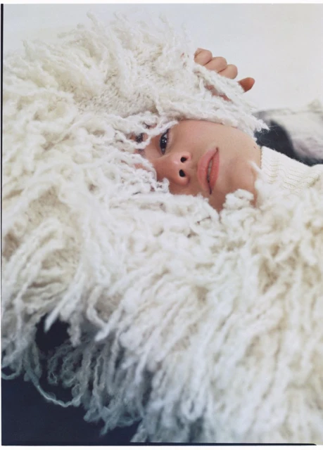 Woman with a neutral expression and soft lighting surrounded by a fluffy white garment or blanket in a portrait-style shot.