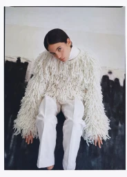 Model seated against a textured wall wearing a textured white fringed sweater and matching trousers.