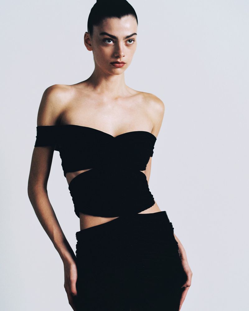 Portrait of a slender woman in black off-shoulder cutout dress against a plain background, looking away with a neutral expression.