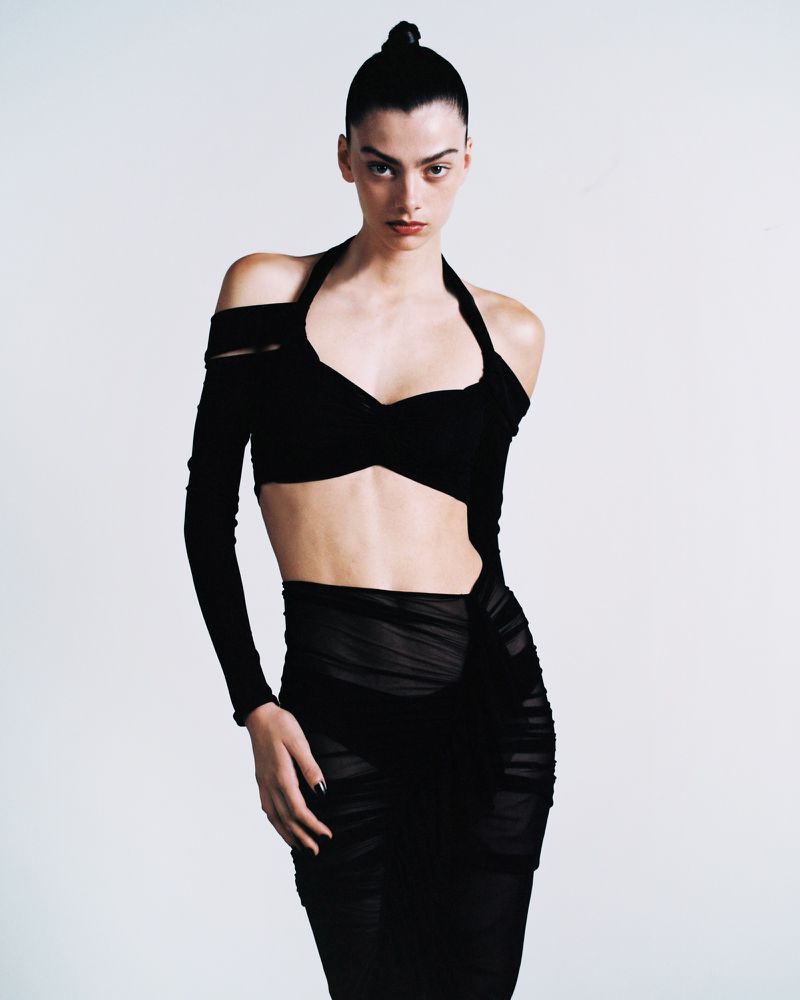 A slender woman with dark hair poses in a black two-piece outfit against a white backdrop in a studio setting, showcasing high-fashion attire.