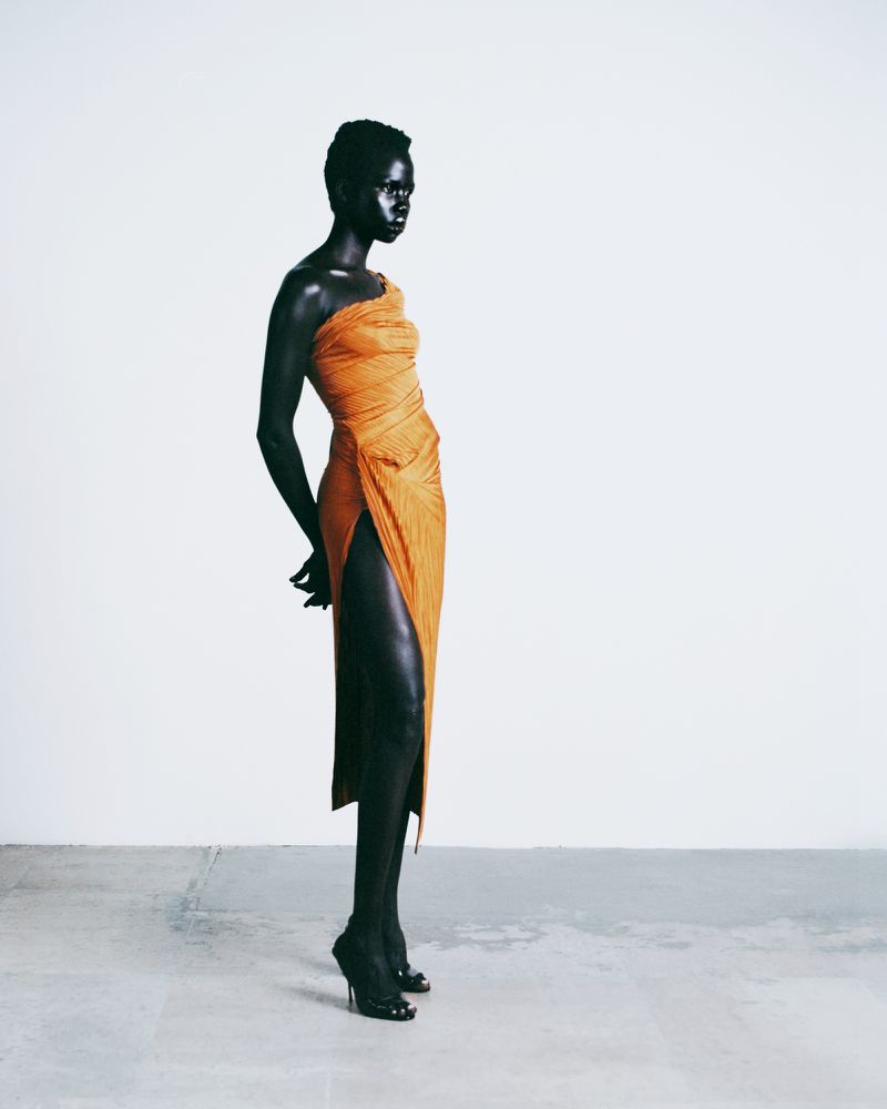 Striking fashion shot of a Black female model in a draped orange gown, posing in a white studio with bright lighting and a minimalist aesthetic.