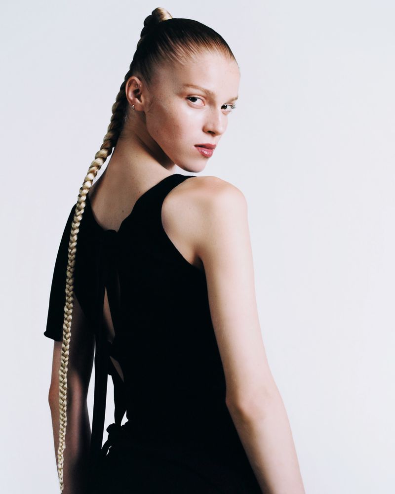 Portrait of a female model with braided ponytail, wearing a black sleeveless dress, standing against a white studio backdrop, looking over shoulder.