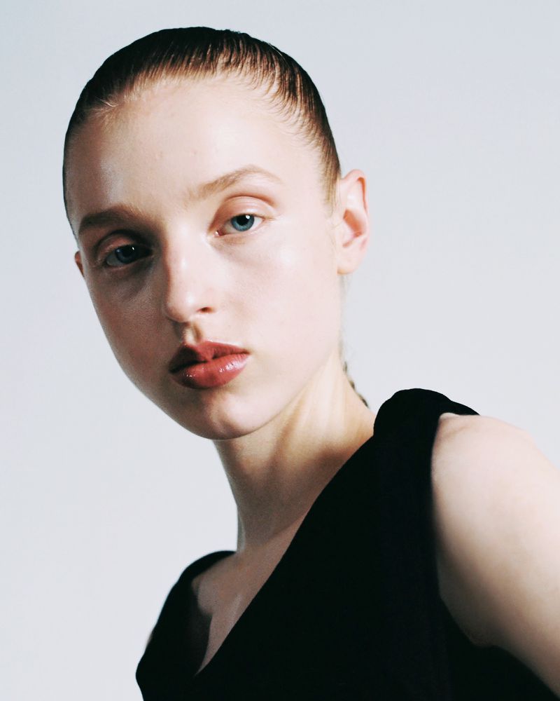 Model with slicked-back hair, neutral makeup, and glossy lips wearing a black sleeveless top against a plain white background.