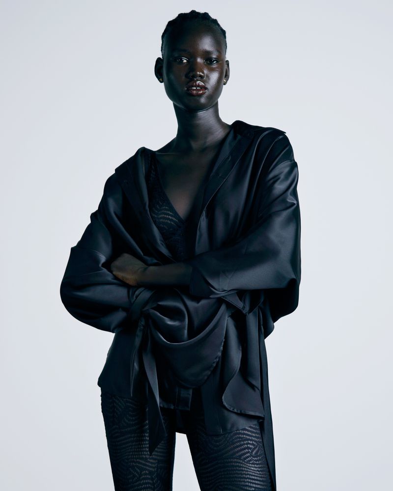 Portrait of a dark-skinned woman in a black silky outfit with arms crossed, standing against a bright white background in a studio.