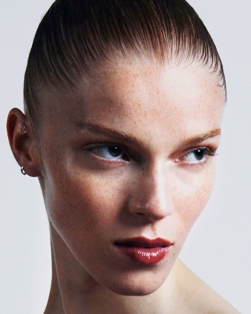 Close-up portrait of a fair-skinned adult female model with freckles, slicked-back hair, and glossy lips against a neutral background.