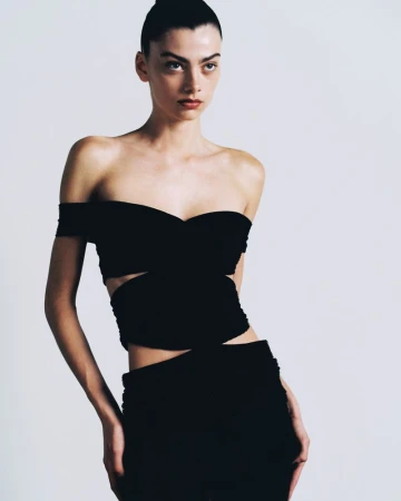 Model with slicked-back dark hair wearing a black off-the-shoulder cutout top and matching black skirt against a pale studio backdrop.