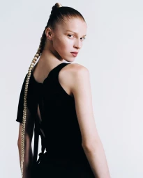 Model with a long blonde braid wearing a black top looking over her shoulder against a white background