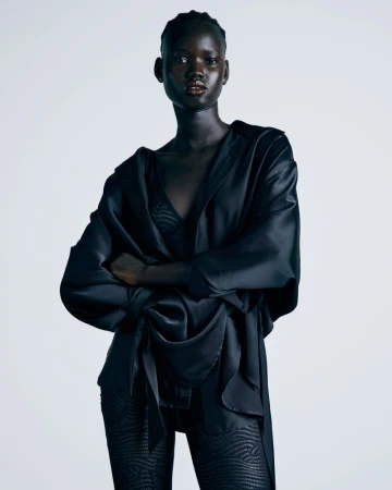 Model posing with crossed arms wearing a draped black satin top and patterned black leggings against a white background