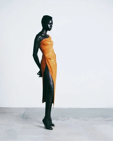 Model standing in an orange draped one-shoulder dress against a pale grey studio background.