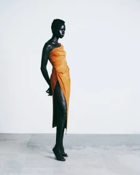Model standing in an orange draped one-shoulder dress against a pale grey studio background.