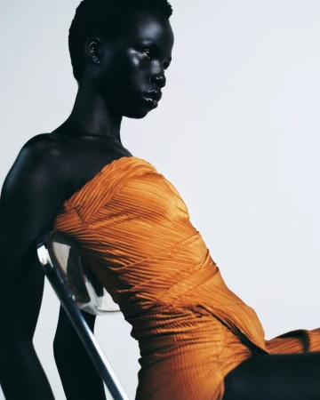 A Black model wearing a pleated orange strapless dress leans back against a clear acrylic chair in front of a white studio backdrop.
