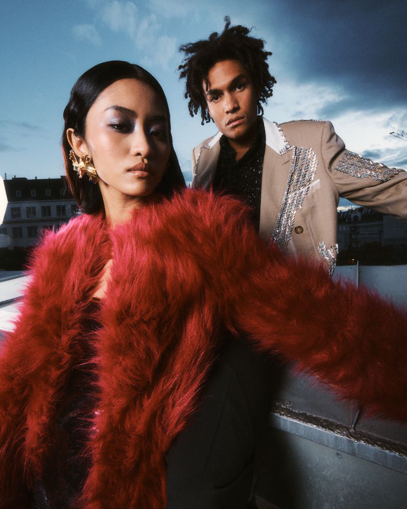 Glamorous fashion portrait of two models in urban outdoor setting; Asian female in red faux fur coat, African-American male in embellished blazer.