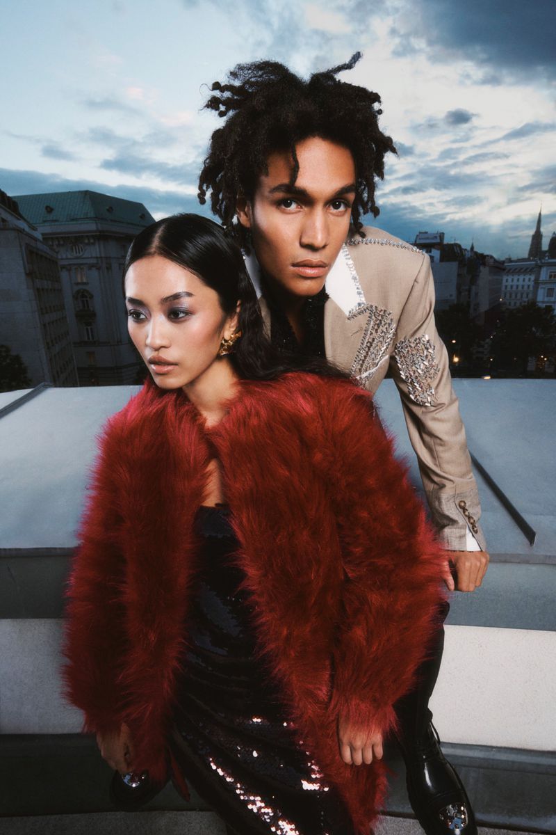 Two fashion models pose on a rooftop against a city skyline, the woman wears a red fur coat, the man is in a cream embellished blazer.