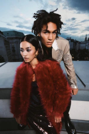 Two fashion models pose on a rooftop against a city skyline, the woman wears a red fur coat, the man is in a cream embellished blazer.