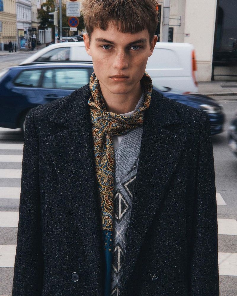 Adult man in a dark wool coat and patterned scarf standing on a crosswalk in an urban street setting with blurred cars and buildings.
