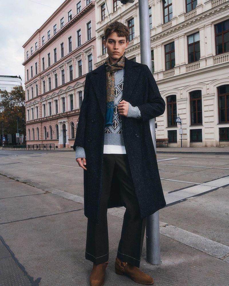 Male fashion model poses outdoors in an urban setting wearing a long navy wool coat, patterned sweater, brown pants, and brown ankle boots.