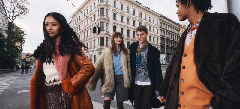Four stylish models in fall and winter apparel, strolling across a city crosswalk, with a backdrop of classic European architecture.