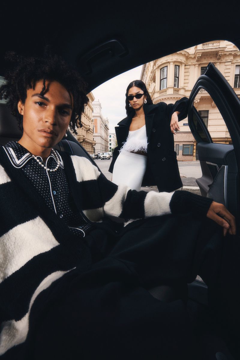 Two fashion models in black and white clothing pose inside a car on a city street, showcasing urban style and modern automotive aesthetics.