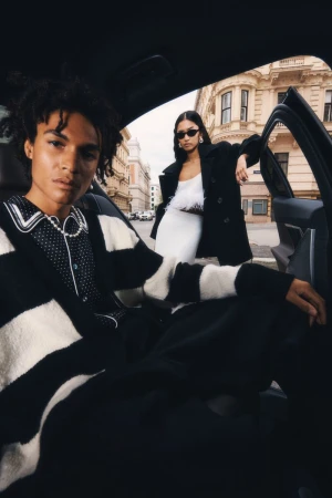 Two fashion models in black and white clothing pose inside a car on a city street, showcasing urban style and modern automotive aesthetics.